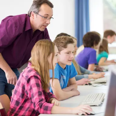 A white teacher leans over a few white adolescents who are using the computer.