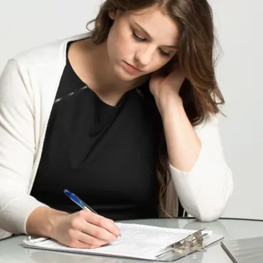 A white woman leans with her elbow on the table while filling out a form on a clipboard. She looks pensive.