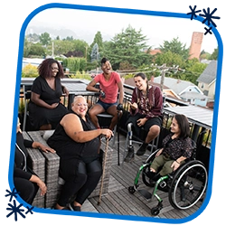 This is a picture in a blue rounded frame with three dark blue asterisks in opposing corners. The picture is an overhead shot of six disabled people of color at a rooftop deck party. An Indigenous Two-Spirit person with a prosthetic leg smiles directly at the camera and gives a thumbs up while everyone else is engaged in conversation.