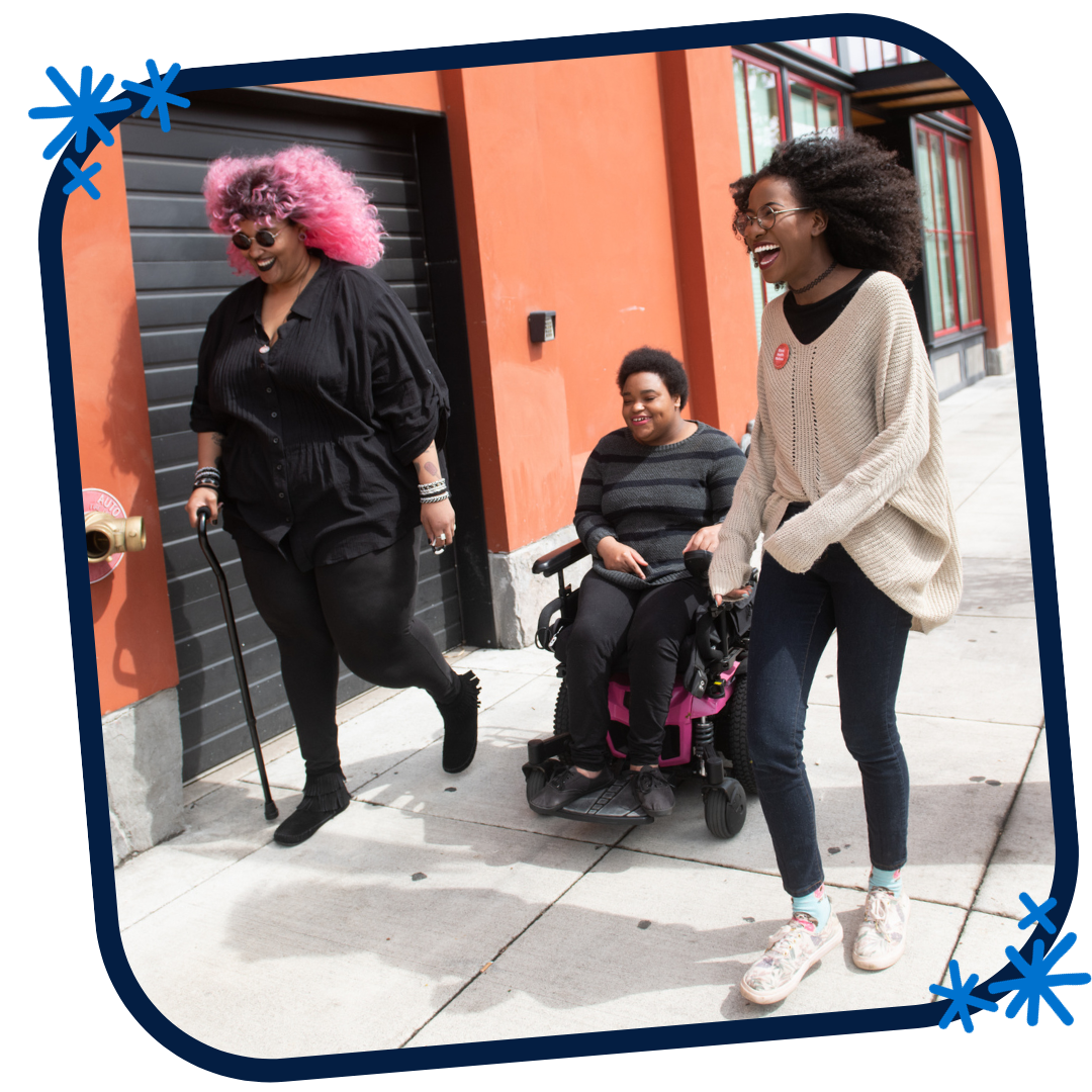 This is a picture in a blue rounded frame with three blue asterisks in opposing corners. This photo features three Black and disabled folx laughing uproariously while strolling down a sidewalk on a windy day. On the left, a non-binary person walks with a cane in one hand and a tangle stim toy in the other. In the middle, a non-binary person rolls along in their power wheelchair. On the right, a femme walks with fabulously windswept hair. A street parking meter is in the background on the right.