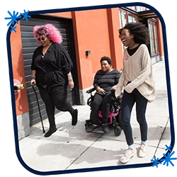 This is a picture in a blue rounded frame with three blue asterisks in opposing corners. This photo features three Black and disabled folx laughing uproariously while strolling down a sidewalk on a windy day. On the left, a non-binary person walks with a cane in one hand and a tangle stim toy in the other. In the middle, a non-binary person rolls along in their power wheelchair. On the right, a femme walks with fabulously windswept hair. A street parking meter is in the background on the right.