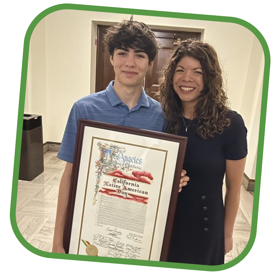 This is a picture of Ravena Soto, on the right wearing black, and her son, on the left wearing a blue shirt. Her son is holding a framed document with the title that reads, “California Native American Day.” 