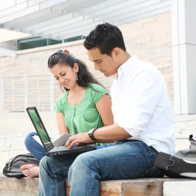 A man using a laptop while a woman reads, sitting next to each other.