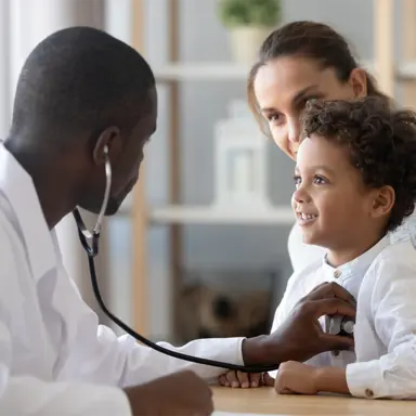A Black male doctor holding a stethoscope to a brown-skinned child's heart. Both the child and his light-skinned mother behind him are smiling.